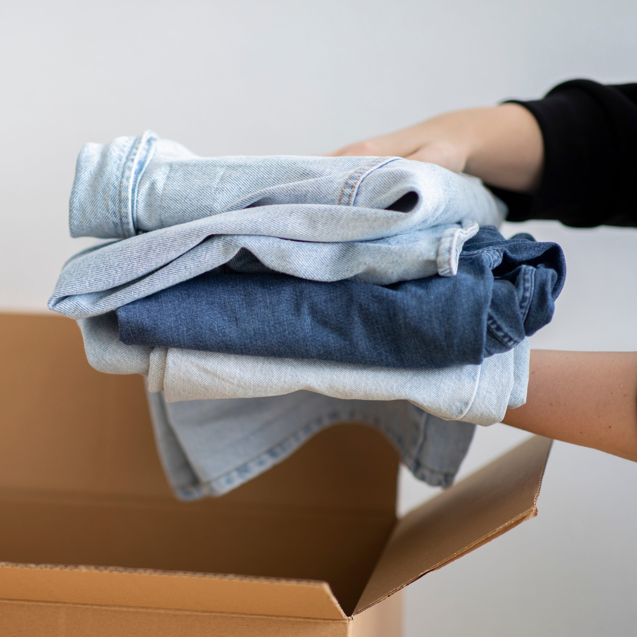 Image of a person's hands holding a pile of folded jeans over a cardboard box.